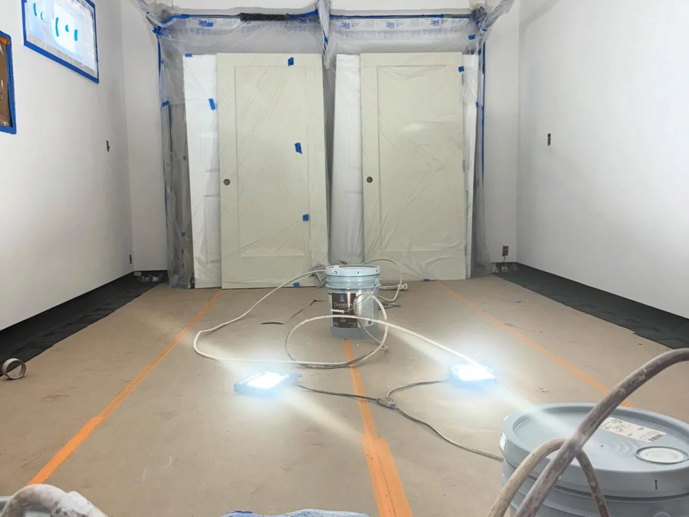 An interior room fully masked with plastic sheeting and tape, floor protection taped down, and work lights on the floor before any paint is opened.